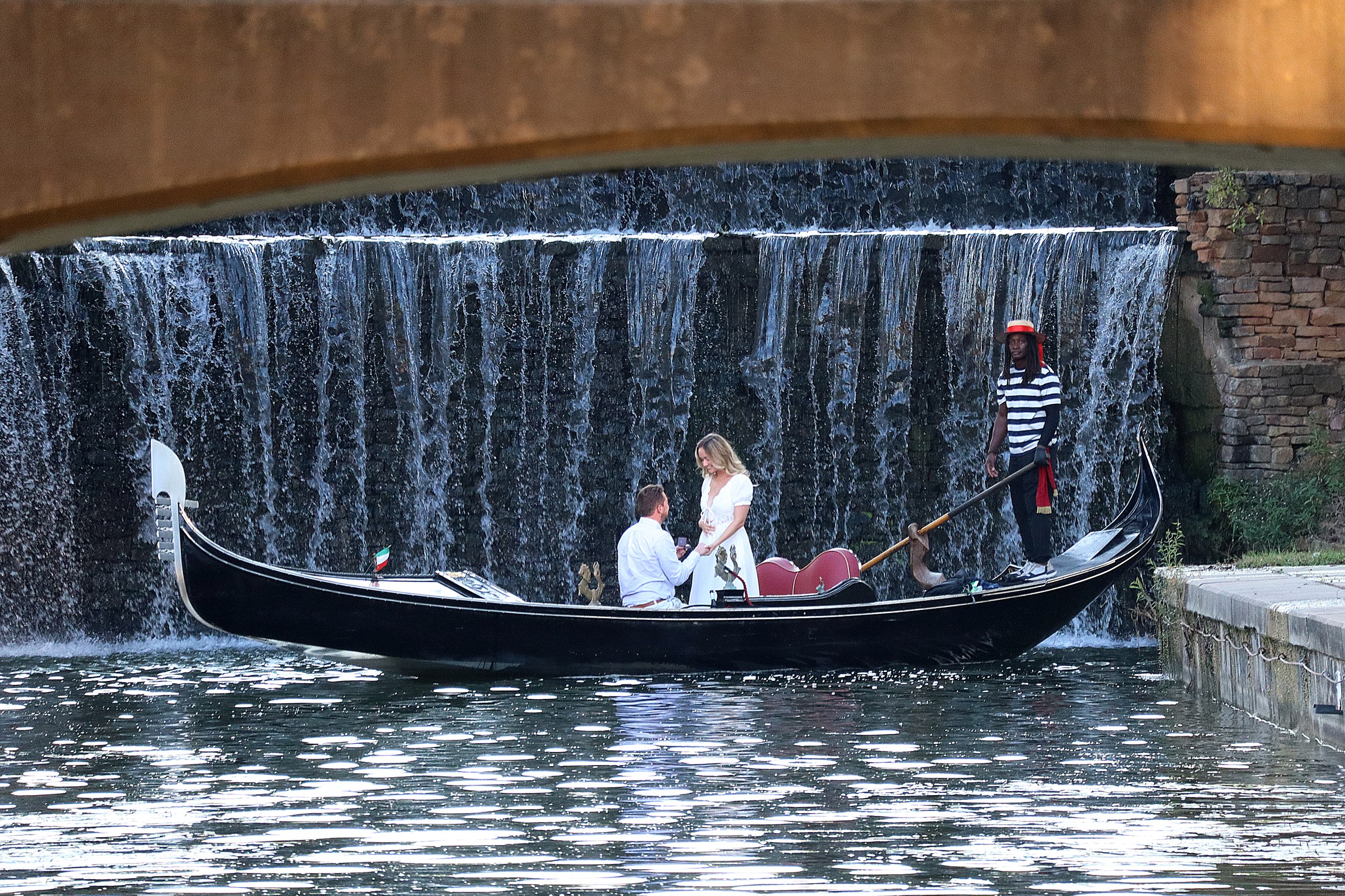 Gondola with three people in front of a waterfall under a bridge.