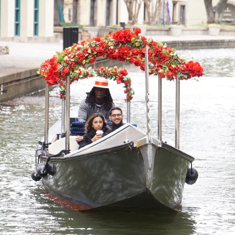 Two people sitting on a boat with a canopy of red flowers on a canal.