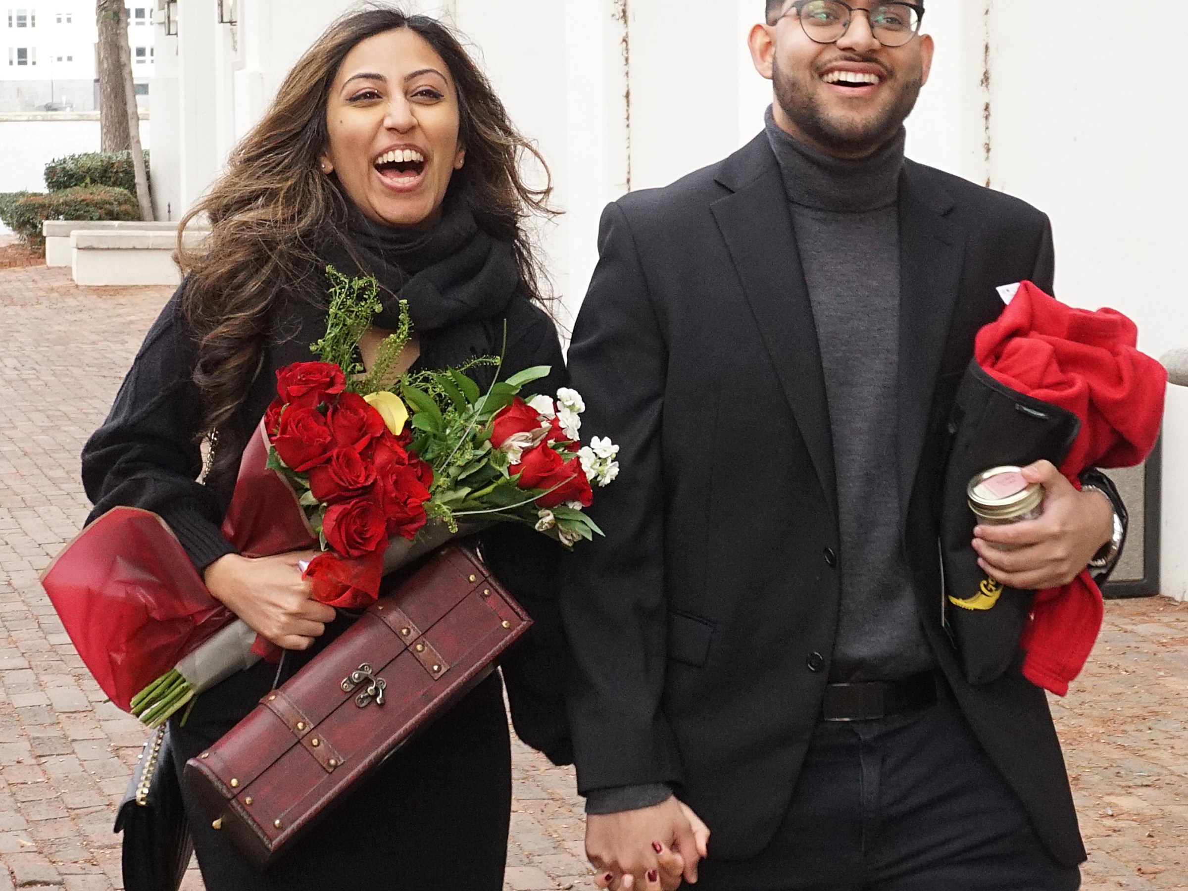 Couple smiling and walking, holding roses and hands, dressed formally.