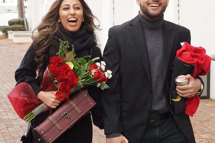 Couple smiling and walking, holding roses and hands, dressed formally.