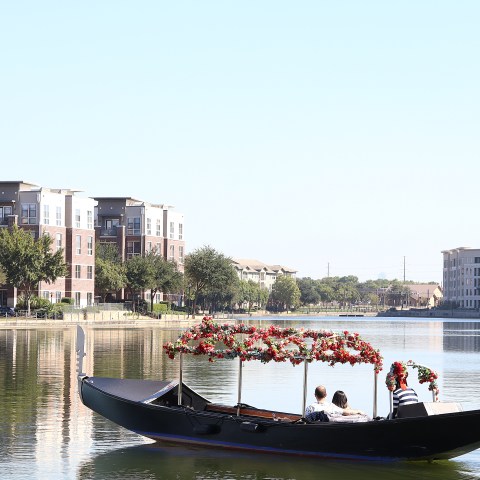 People in a decorated gondola on a calm lake near waterfront buildings.