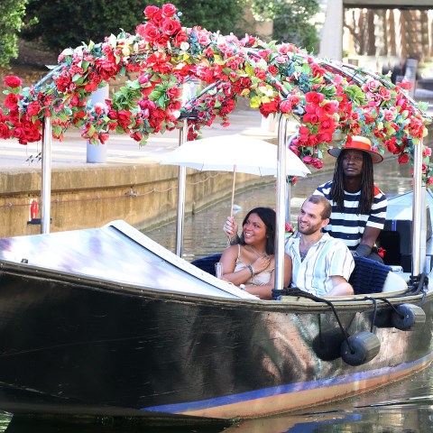 Three people on a boat with a canopy of red flowers on a canal.