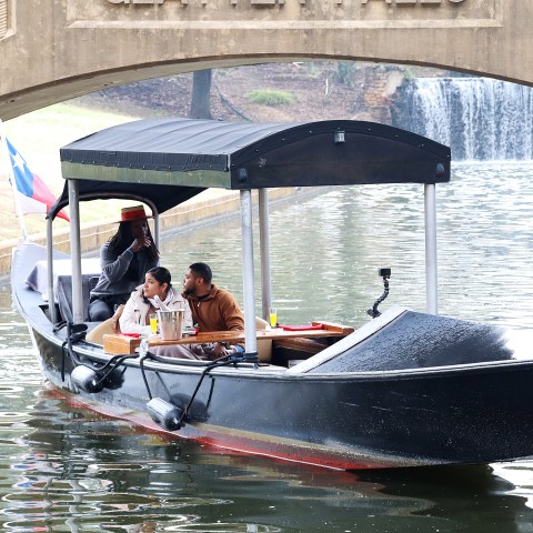 People on a covered boat with flags, passing under a bridge.